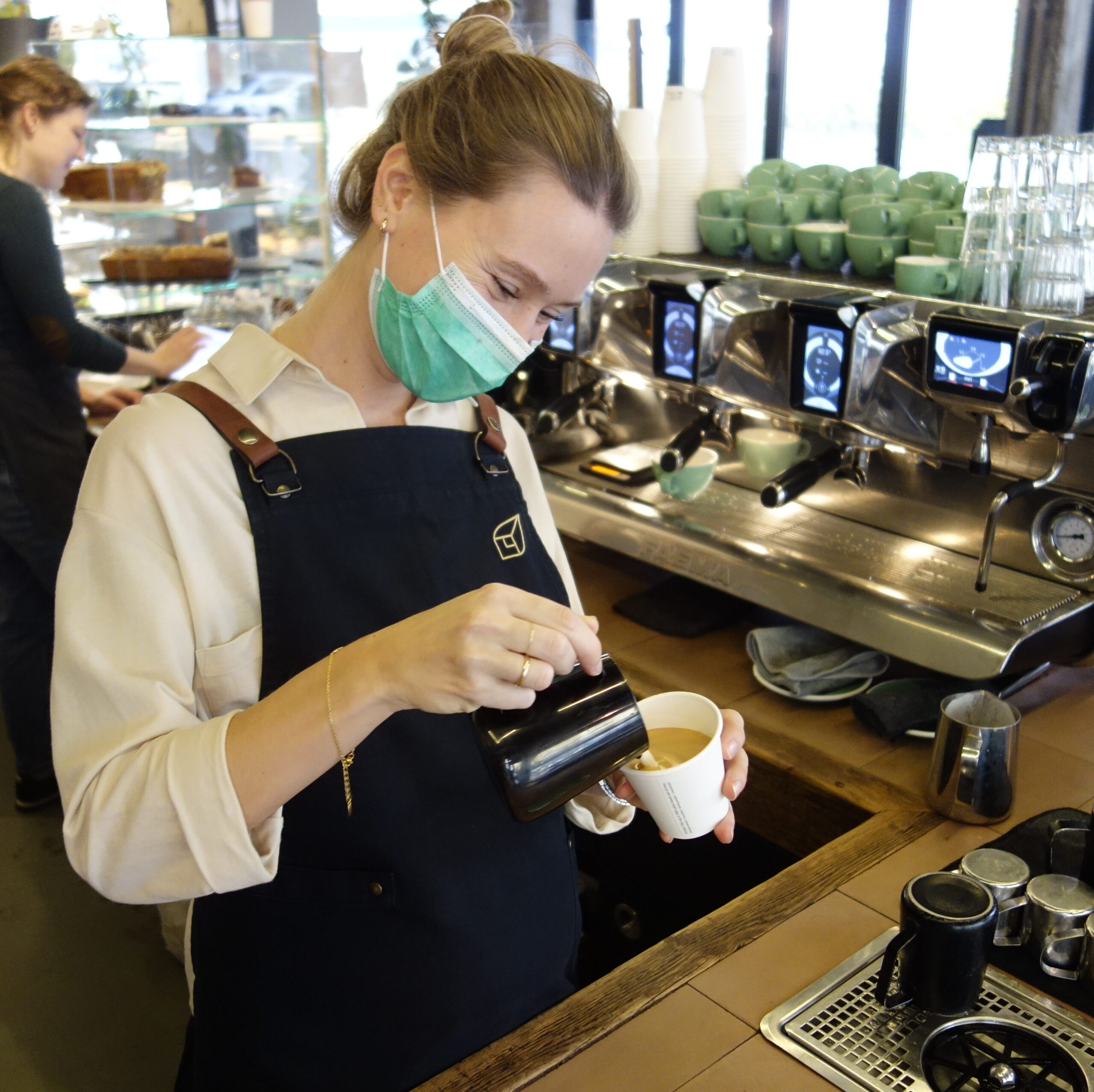 Cafe- Mitarbeiterin mit Barista Schürze macht Cappuccino hinter Kaffeemaschine